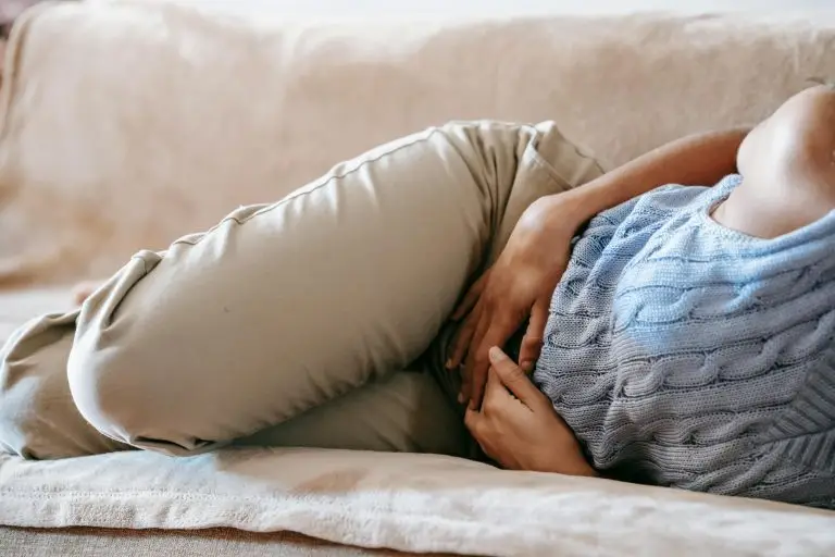 Person lying on a couch with both hands on their abdomen, reflecting the stillness and fatigue that often accompany persistent pain.